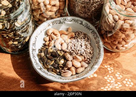 Noccioline, pistacchi, semi di girasole e semi di zucca in vasetti di vetro e ciotole di porcellana sul tavolo. Spuntini sani Foto Stock