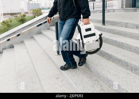 Uomo che porta un monociclo elettrico sulla strada della città. Veicolo portatile portatile per trasporto individuale. Uomo su monoruota elettrica guida veloce (EUC) al lavoro Foto Stock