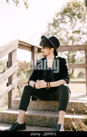 Il giovane uomo in cappello nero siede su gradini di pietra con ringhiera di legno Foto Stock