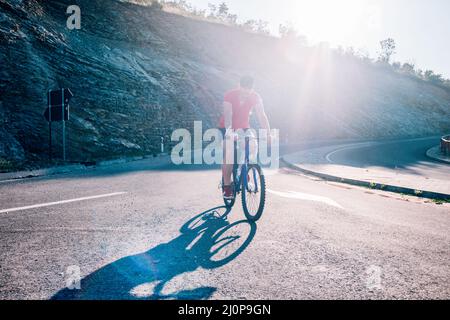 Montare maschio ciclista biker in sella alla sua moto ciclo su una strada asfaltata al tramonto mentre il sole tramonta attraverso la sua ruota. Foto Stock