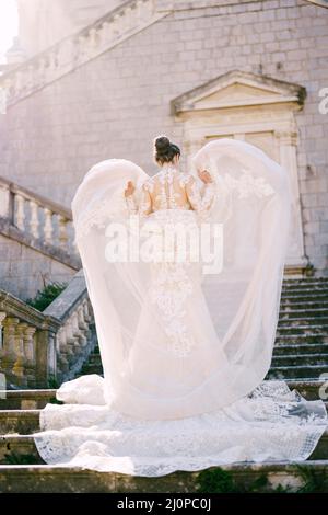 La sposa sta in piedi sui gradini di pietra con le mani che sollevano lo strato superiore del suo vestito Foto Stock