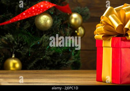 Regalo di Natale in una scatola quadrata con un grande arco d'oro si trova sul tavolo. Sfondo di un albero di Natale verde con palline d'oro, ho Foto Stock