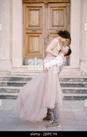 Lo sposo tiene la sposa nelle sue braccia di fronte all'ingresso dell'antico edificio Foto Stock