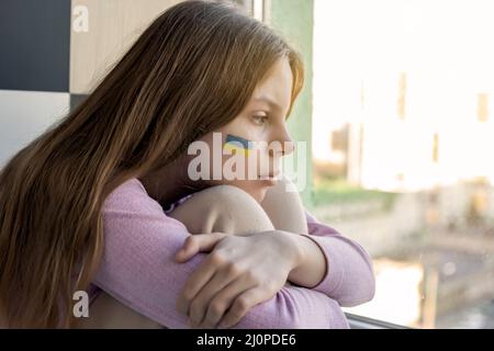 Una triste ragazza Ucraina con una bandiera Ucraina dipinta sulla sua guancia si siede e guarda fuori dalla finestra, in attesa della fine della guerra. Crisi, guerra, nessuna pace Foto Stock