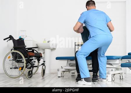Fisioterapista maschile che aiuta un paziente con una disabilità che utilizza una sedia a rotelle, per alzarsi in ospedale di riabilitazione. Foto Stock