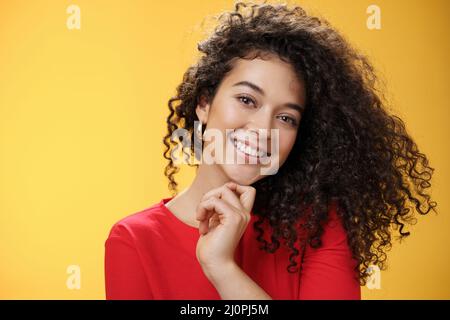 Primo piano di elegante e allegra donna dai capelli ricci in abito rosso testa inclinabile che tocca sensualmente il mento con le dita e. Foto Stock