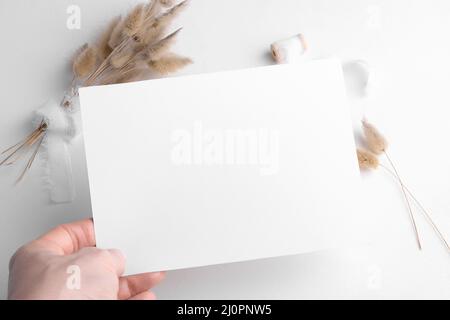 Mockup carta invito cancelleria di nozze 7x5 su sfondo bianco con arredamento boho, femmina mano tenere vuota carta mockup. Minimal bohémien bianco mockup, Foto Stock