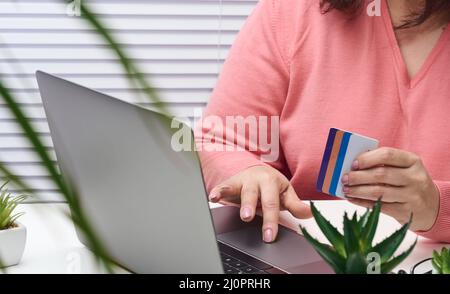 Donna in un maglione rosa effettua acquisti online con un laptop, una carta di credito in mano. Donna seduta ad un tavolo bianco e guardando Foto Stock