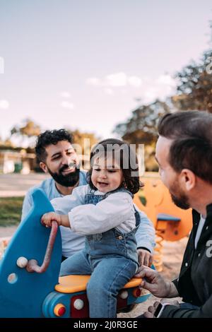 Coppia gay maschile che gioca con la loro figlia su una seesaw nel parco. Moderno concetto di famiglia. Verticale Foto Stock