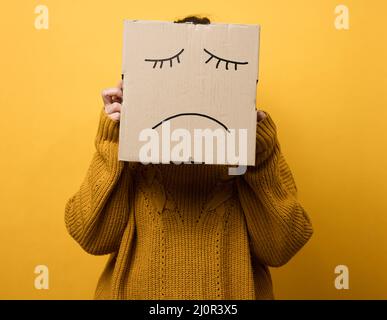 Una donna in un maglione marrone con una scatola sulla testa si erge su uno sfondo giallo. Triste concetto di grimace, stress e depressione. Uomini Foto Stock