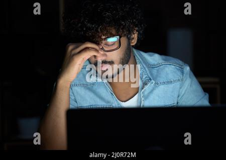 Stanco sonnolento giovane uomo indiano che indossa occhiali utilizzando il computer che funziona tardi. Foto Stock
