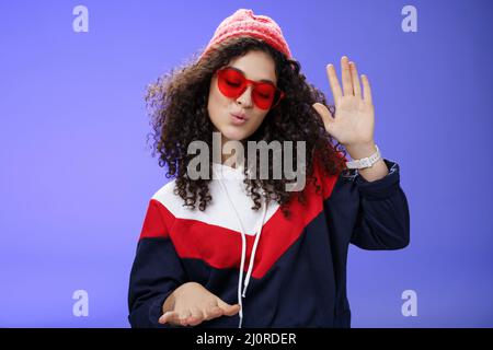 Studio girato di dj ragazza cool ed elegante in rosso beanie e occhiali da sole che alzano la palma come godere di musica fredda e ballare al ritmo f Foto Stock