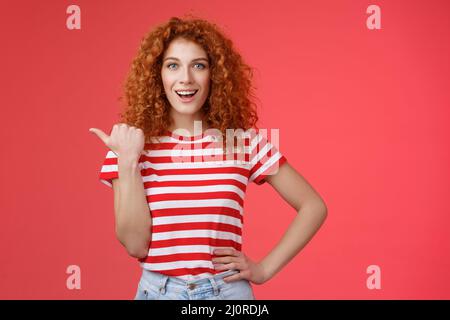 Sassy buon-looking emotivo felice sorridente rosso europeo femmina riccio acconciatura puntando pollice sinistra grin assertivo cheeky tenere Foto Stock