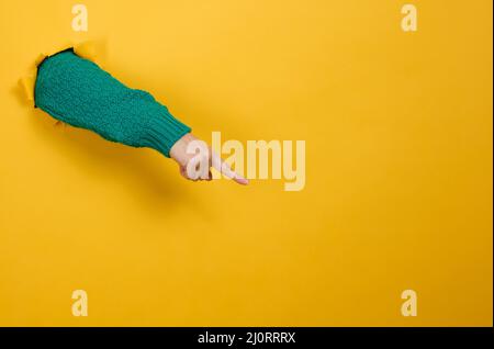 La mano femmina sporge da un foro strappato su uno sfondo di carta giallo, parte del corpo punta con il dito indice a lato. C Foto Stock