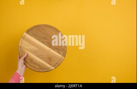 Donna che tiene in mano un piano di pizza vuoto rotondo di legno, parte del corpo su sfondo giallo Foto Stock