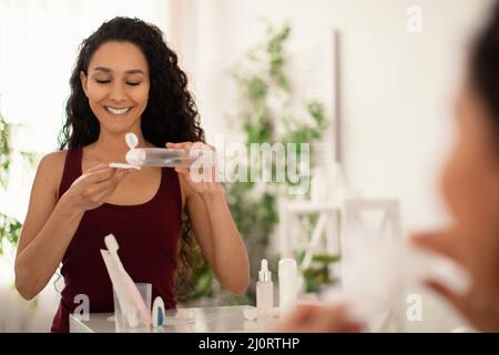 Bella giovane donna che rimuove il trucco con lozione o tonico, utilizzando il batuffolo di cotone di fronte allo specchio in bagno, spazio vuoto Foto Stock