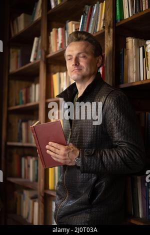 Bell'uomo aristocratico dalle delicate caratteristiche si erige in una Libreria con un vecchio libro rosso nelle sue mani, gli occhi chiusi con Pl Foto Stock