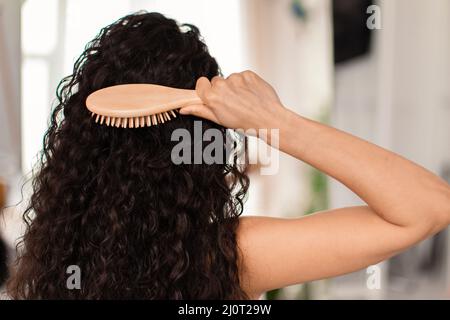 Vista posteriore della giovane donna bruna spazzolando i suoi lunghi capelli ondulati con spazzola di legno a casa Foto Stock