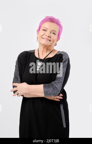 Sorridente vecchia donna con corti capelli rosa tiene le mani incrociate. Concetto di emozione positiva. Foto Stock