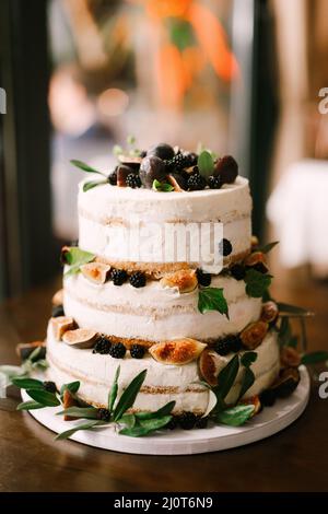 Torta nuziale a tre piani decorata con more, fichi, prugne e foglie verdi si erge sul tavolo Foto Stock