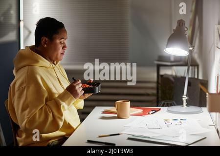 Ritratto laterale di donna latino-americana che mangia takeout alla scrivania mentre lavora a tarda notte in ufficio, spazio copia Foto Stock