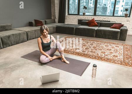 Donna sportiva si siede su un mat sportivo e ha una Video Chat online con la sua allenatore di fitness sul laptop durante il periodo di quarantena. Hom Foto Stock