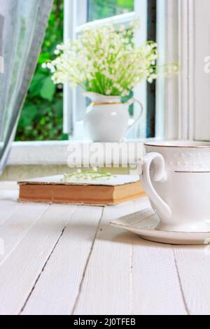 Una tazza di tè o caffè e un libro su un tavolo retrò in legno bianco e un bouquet di Lily della valle fiori sul vento Foto Stock