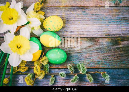 Composizione pasquale di narcissus fiori in fiore rametti di salice e uova di Pasqua con un motivo di colore giallo e verde su un bosco Foto Stock