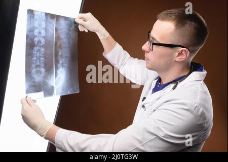 Il radiologo medico giovane in occhiali e vestiti medici guarda l'immagine radiografica lumen della spina dorsale su uno sfondo marrone. Foto Stock