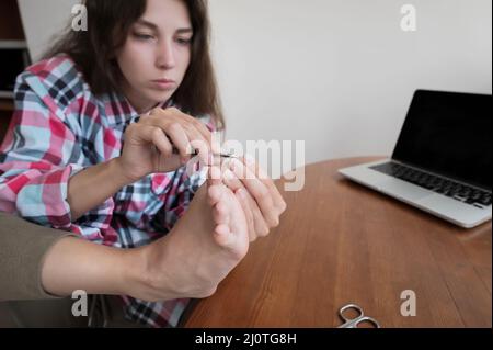 Attraente giovane donna caucasica in una camicia seduta in cucina facendosi un pedicure. Cura della casa interna Foto Stock