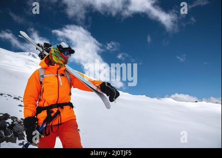 Ritratto di montagna di uno sciatore freeride professionista in abbigliamento arancione con bastoncini da sci e sci sulle spalle. Si alza in altezza in t Foto Stock