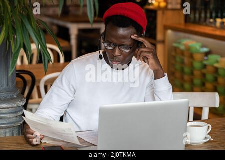 Hipster uomo afro-americano pensivo in occhiali di distanza lavorando su laptop con documenti in caffè Foto Stock
