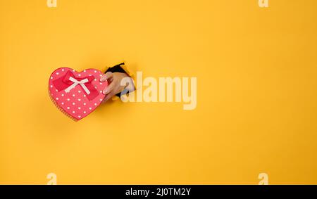 La mano femmina tiene una scatola rossa con un regalo su uno sfondo giallo, parte del corpo sporge da un buco strappato in uno sfondo di carta Foto Stock