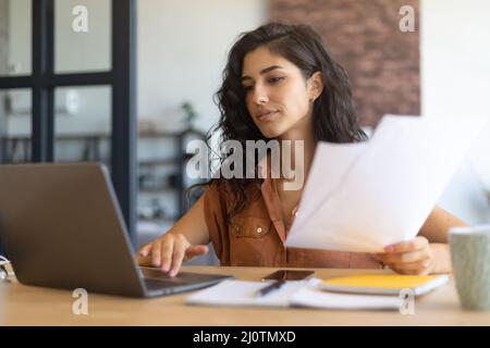 Giovane donna d'affari focalizzata che lavora o studia su un notebook, tiene documenti, si siede alla scrivania all'interno, spazio per le copie Foto Stock