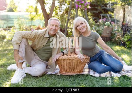 Felice coppia sposata anziana che ha picnic all'aperto, seduta su coperta e sorridente alla macchina fotografica, godendo la sera calda Foto Stock