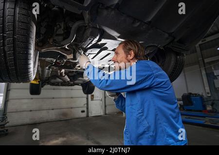 Meccanico in abiti da lavoro blu e guanti protettivi in prossimità di una macchina sollevata, guardando verso l'alto e ispezionando le leve Foto Stock