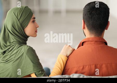 Moglie mediorientale infelice che tocca la spalla del marito seduta a casa Foto Stock