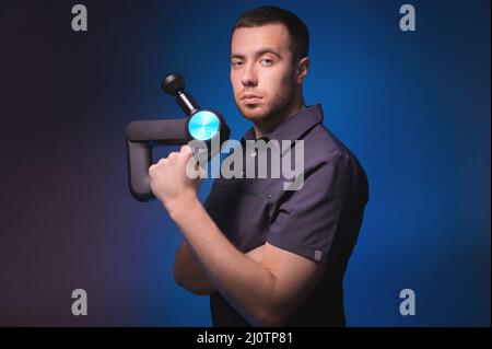 Ritratto di un massaggiatore professionista sicuro con un massaggiatore a percussione nelle sue mani. Tasto basso, sfondo blu. Onda d'urto massa Foto Stock