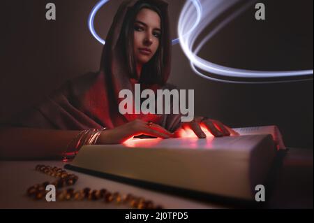 Giovane caucasico attraente fortuna teller strega astrologo donna in una felpa con cappuccio scuro con palme luminose legge il libro Foto Stock