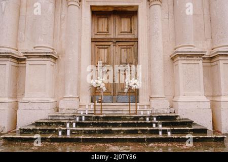 Mazzi di fiori si eriscono su plinti dorati sui gradini bagnati di fronte ad una porta di legno della chiesa Foto Stock