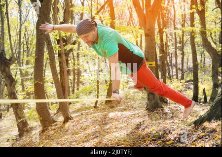 Un uomo barbuto di età fa un trucco sul suo braccio che si bilancia su un lento slackline nella foresta d'autunno. Il concetto di vita attiva Foto Stock