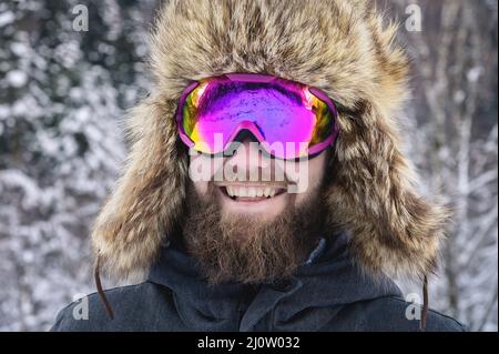 Primo piano ritratto di un felice sciatore bearded snowboarder in una maschera da sci con occhiali e un grande cappello di pelliccia vecchia scuola su sfondo o Foto Stock