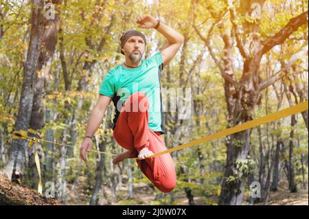 A bearded man aged balancing on a slackline in the autumn forest on a sunny day. The concept of sports leisure at the age of for Stock Photo