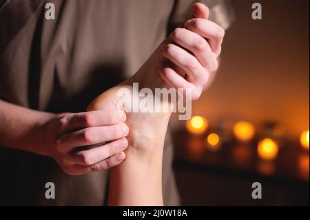 Primo piano di un massaggiatore maschile che fa un massaggio gambe e piedi in una stanza oscura sullo sfondo di candele massaggio professionale in Foto Stock