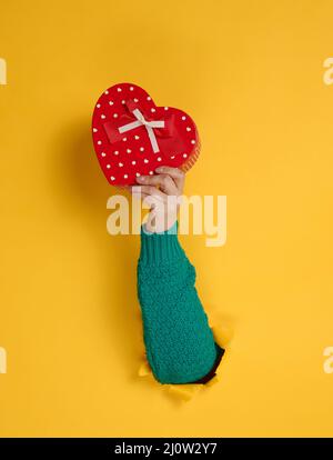 La mano femmina tiene una scatola rossa con un regalo su uno sfondo giallo, parte del corpo sporge da un buco strappato in uno sfondo di carta Foto Stock