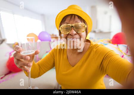 Donna anziana in occhiali a serranda per videochiamare mentre si gusta lo champagne durante la festa a casa Foto Stock