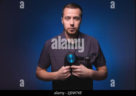 Ritratto di un massaggiatore professionista sicuro con un massaggiatore a percussione nelle sue mani. Tasto basso, sfondo blu. Onda d'urto massa Foto Stock