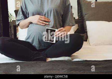 Scatto corto della donna incinta che tiene l'ultrasuono di gravidanza o il sonogramma del bambino mentre si siede sul letto Foto Stock