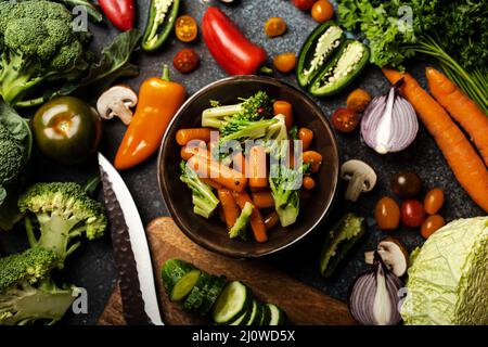Insalata di broccoli di carota al vapore in ciotola e verdure fresche tagliate assortite su tavola rustica in cemento Foto Stock