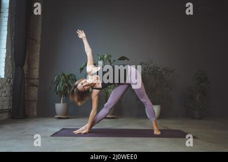 Donna solitaria in Sportswear che fa il fianco in un appartamento in una metropoli. Sport Fit Woman Practices Hatha Yoga. Lunghezza intera. Grigio Foto Stock